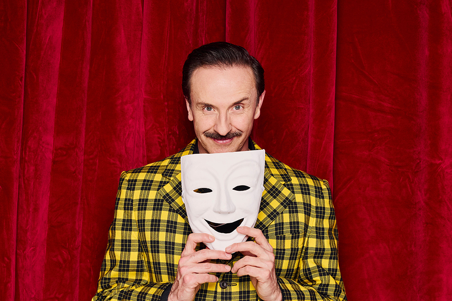 Man wears yellow and black check suit holding a theatre mask standing in front of a red stage curtain