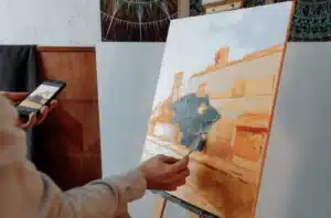 A person paints with one hand while using a cell phone in the other, illustrating the blend of art and technology.