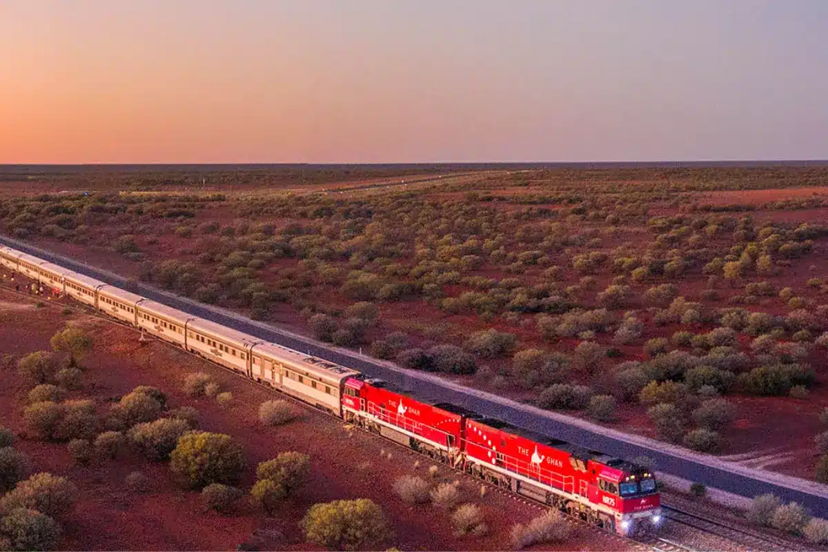 A red train traverses the desert landscape, illuminated by the warm hues of a sunset in the background.