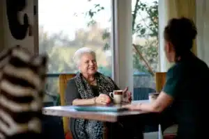 An older woman sits at a table, enjoying a cup of coffee with a warm smile on her face.