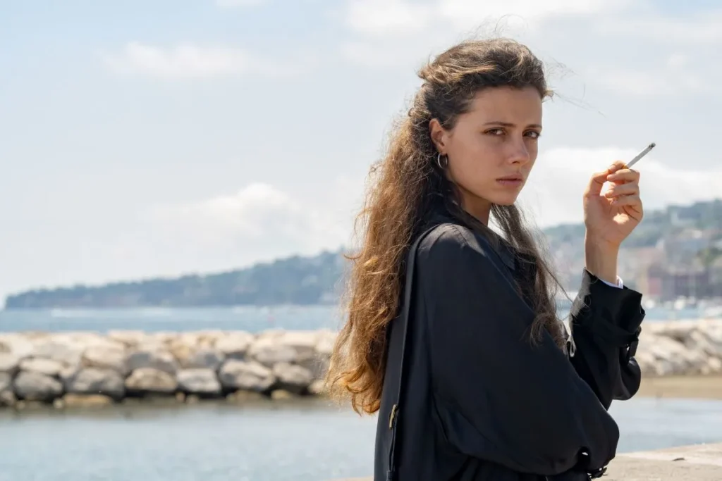 Woman with long hair standing by the water, holding a cigarette, looking pensively at the camera.