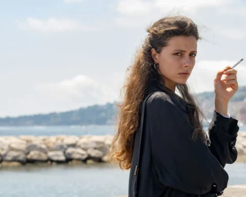 Woman with long hair standing by the water, holding a cigarette, looking pensively at the camera.