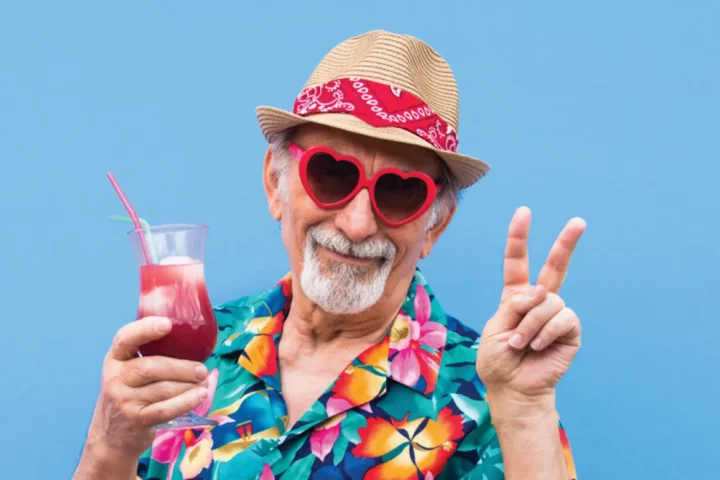 An elderly man in a tropical shirt and sunglasses enjoys a refreshing drink, smiling in a sunny setting.