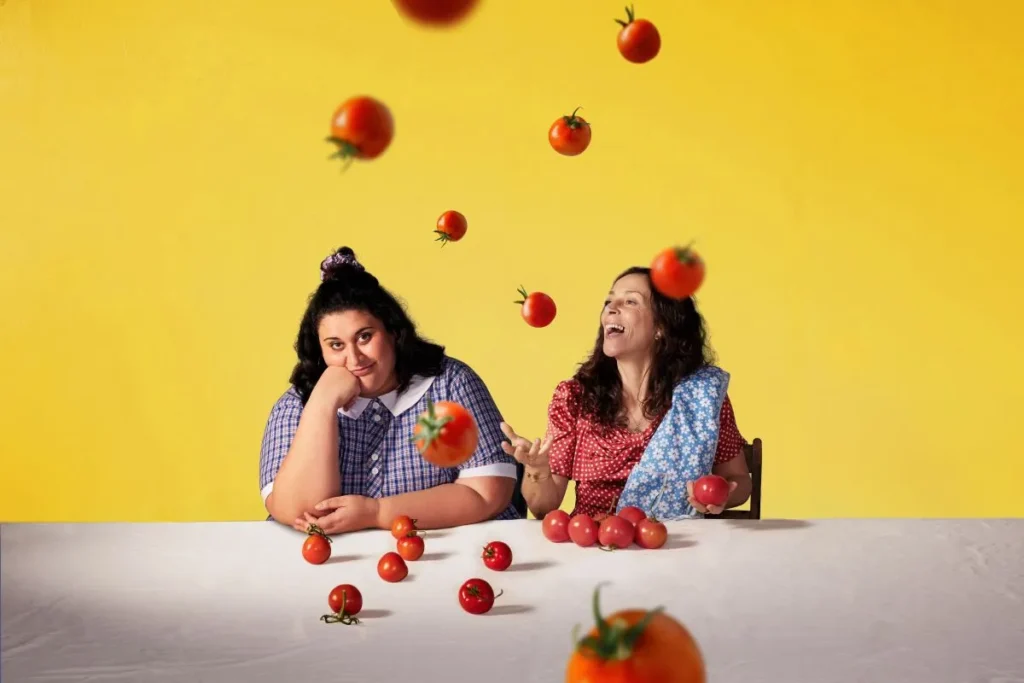 Two women sit at a table, laughing as tomatoes fall from the ceiling around them.