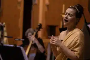 A woman in a yellow dress passionately sings in front of a full orchestra, captivating the audience with her performance.