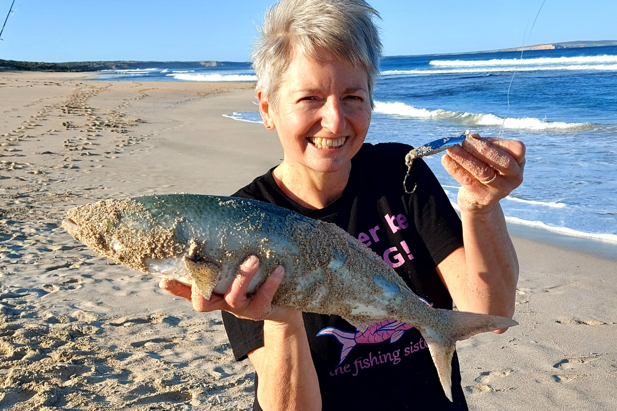 A woman stands on the beach, proudly holding a freshly caught fish in her hands, with waves gently lapping at the shore.