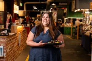 A woman stands in a market, proudly holding a plate of colorful food, showcasing local cuisine and vibrant ingredients.