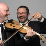 Two men in formal suits and ties are holding a violin, showcasing a blend of elegance and musical artistry.