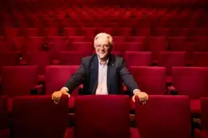 A man with crossed arms sits in a red auditorium, surrounded by plush red seats and a vibrant atmosphere.