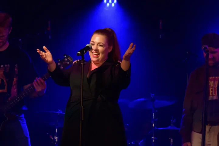 A woman passionately singing on stage, surrounded by fellow musicians, creating a vibrant musical atmosphere.