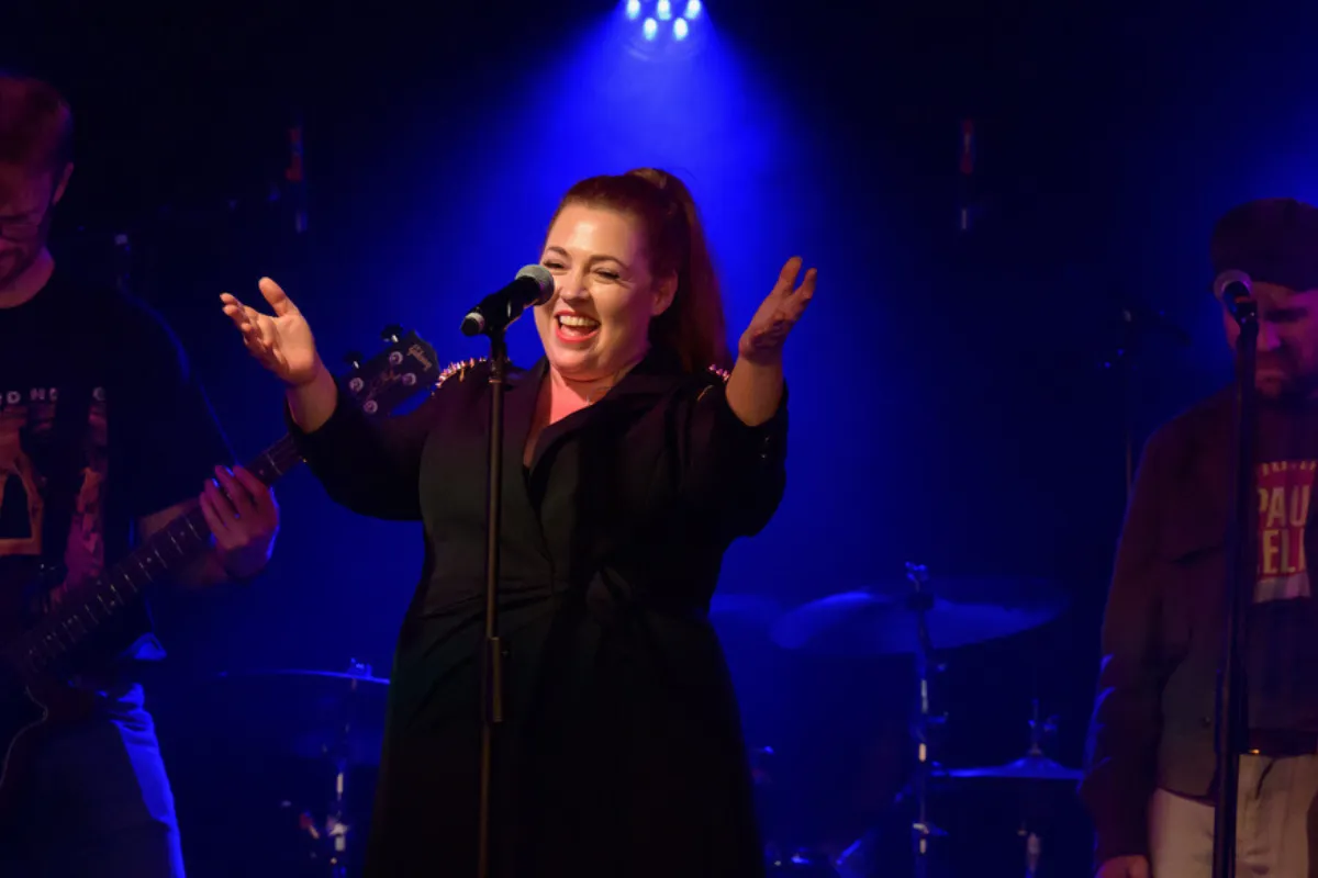 A woman passionately singing on stage, surrounded by fellow musicians, creating a vibrant musical atmosphere.