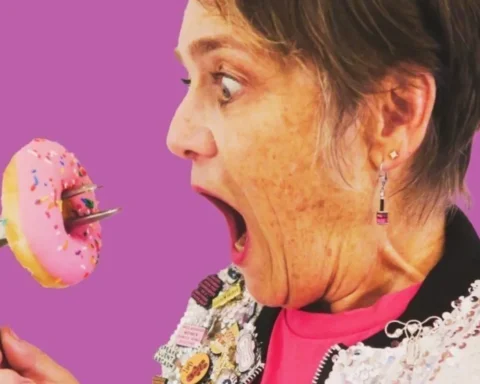 A woman holds a pink doughnut in one hand while gripping a nail gun in the other, showcasing a quirky contrast.