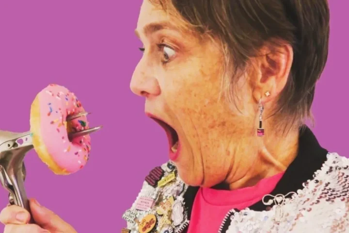 A woman holds a pink doughnut in one hand while gripping a nail gun in the other, showcasing a quirky contrast.