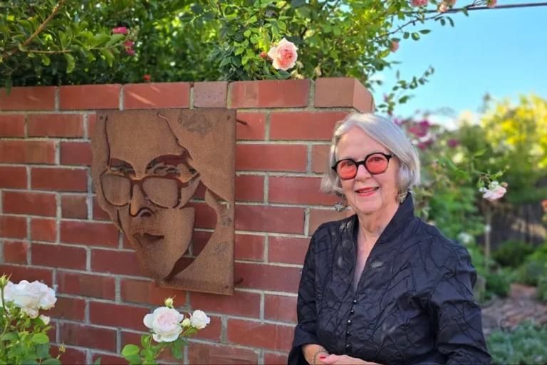 An older woman stands beside a brick wall featuring a painted face, creating an intriguing urban scene.