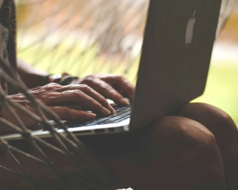 A woman relaxes in a hammock, working on her laptop under a sunny sky.