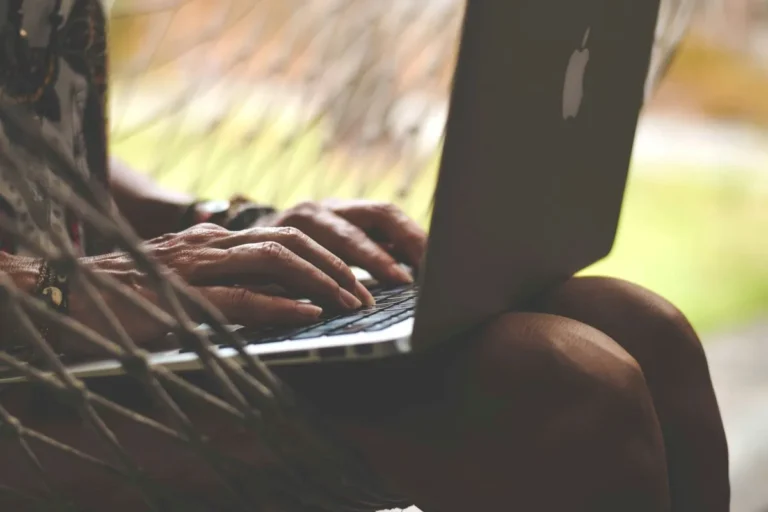 A woman relaxes in a hammock, working on her laptop under a sunny sky.