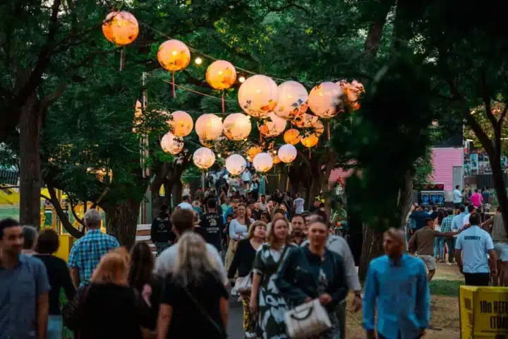 A crowd strolls beneath a tree adorned with colorful paper lanterns, creating a festive and vibrant atmosphere.