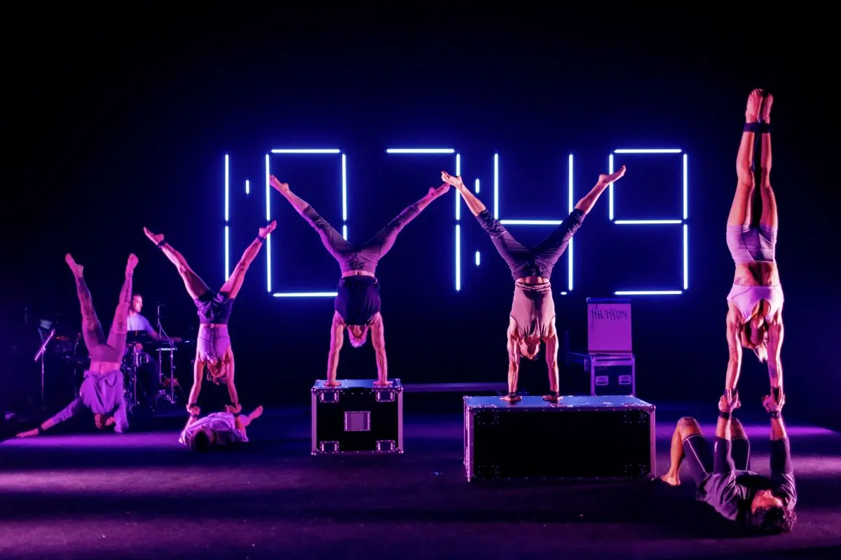 A group of acrobats performing on stage, showcasing their skills with a large clock in the background.