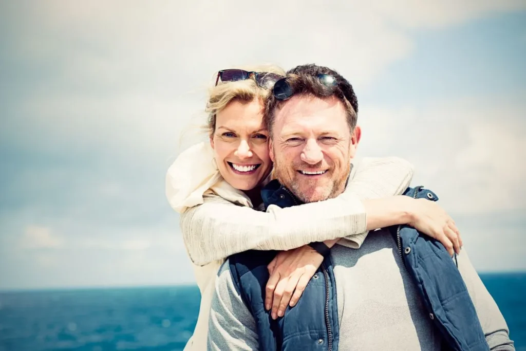 A man and woman embrace warmly on a sandy beach, with waves gently lapping at the shore in the background.