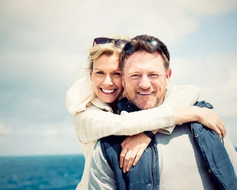 A man and woman embrace warmly on a sandy beach, with waves gently lapping at the shore in the background.