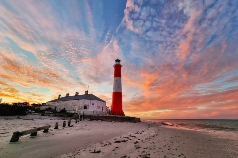 A lighthouse stands tall on a beach, silhouetted against a vibrant sunset sky.