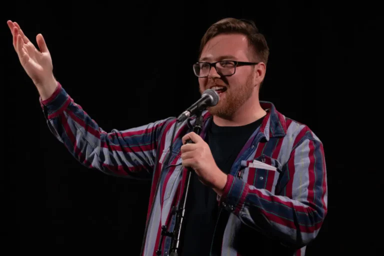 A man wearing glasses and a plaid shirt holds a microphone, appearing engaged in a speaking or performance activity.