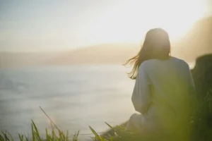 A woman sits on a rock, gazing out at the vast ocean under a clear blue sky.