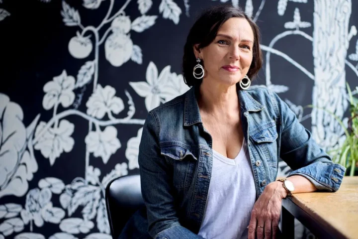 A woman sitting at a table in front of a black and white wall, focused on her work with a thoughtful expression.