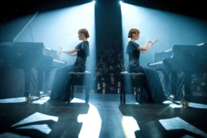 Two women performing a piano duet on stage in front of an engaged audience.