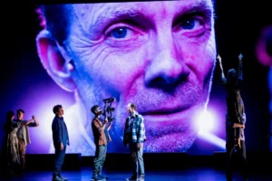 A group of people stands on stage in front of a large screen displaying a man.