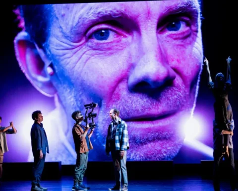 A group of people stands on stage in front of a large screen displaying a man.