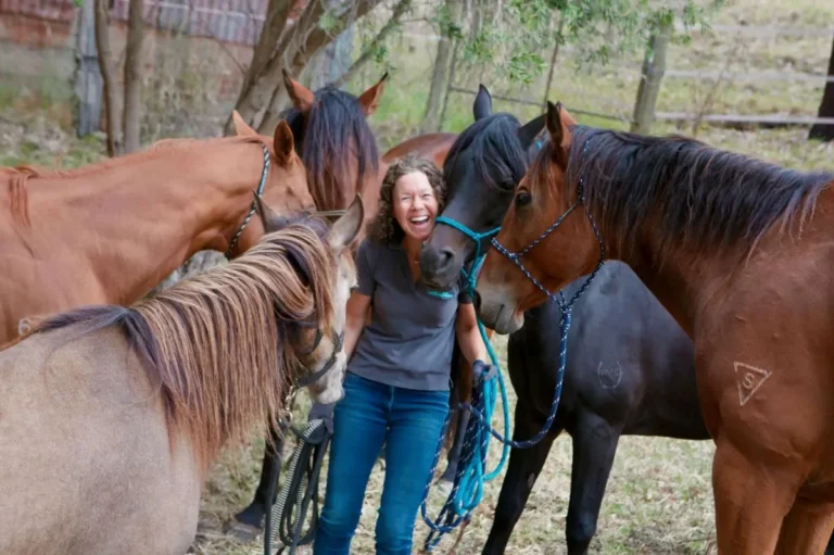 A woman stands among a group of horses in a sunny field, smiling and gently reaching out to one of the horses.