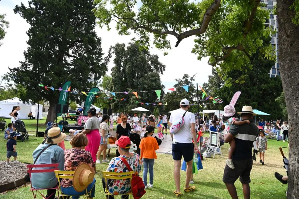 A lively park scene with a crowd of people seated on chairs, surrounded by greenery and enjoying the day.